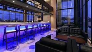 A modern bar area with high stools and a bar counter, large windows, and a seating area with sofas and a coffee table. - Harper Cikarang by ASTON (Cikarang)
