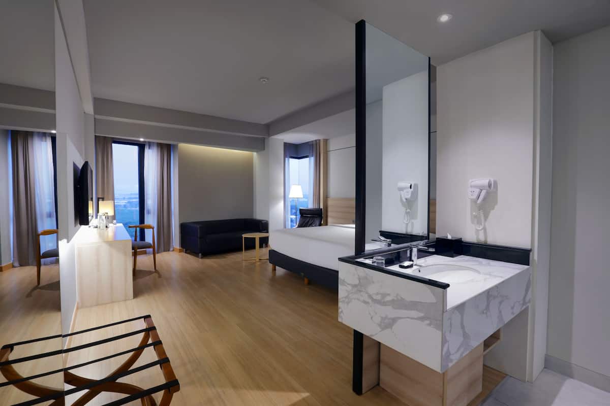 Una habitación de hotel moderna con una cama grande, un área de descanso con vistas, un comedor y un baño con un espejo amplio y un secador de pelo.