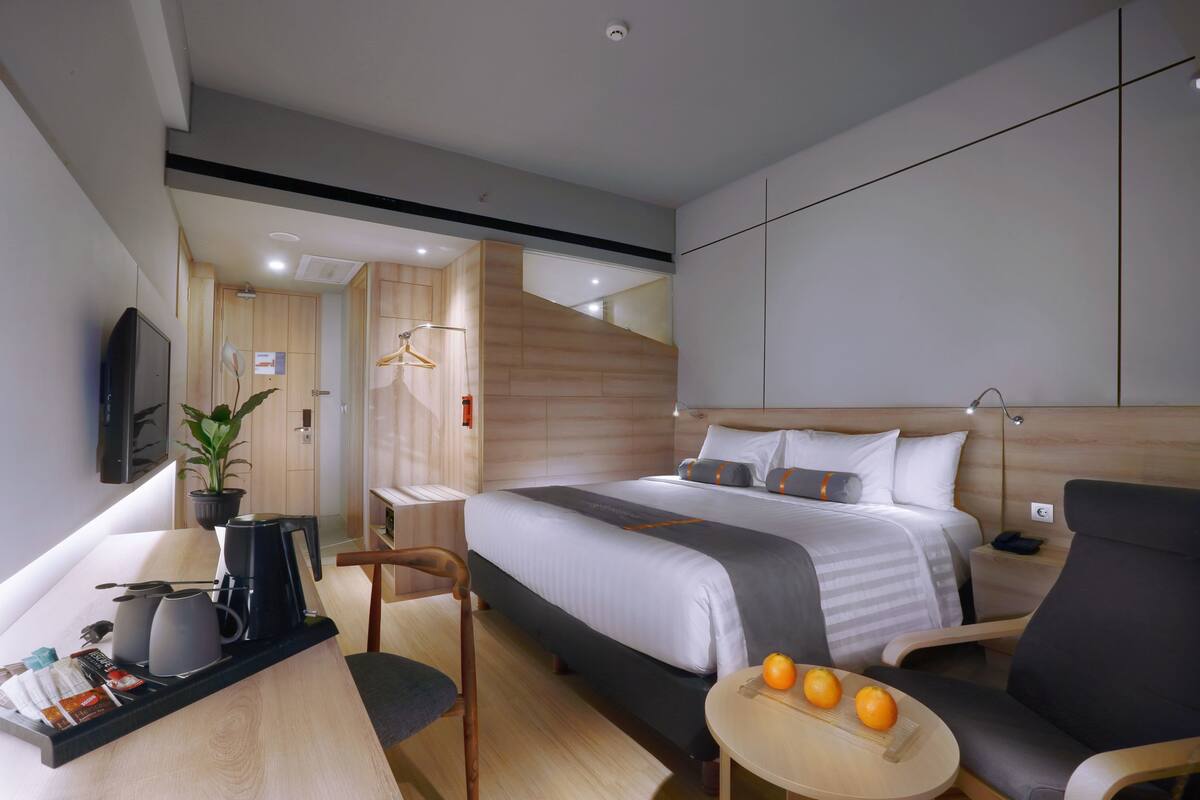 Una habitación de hotel moderna con una cama grande, un escritorio con cafetera, una silla, una mesita con naranjas y un televisor montado en la pared.