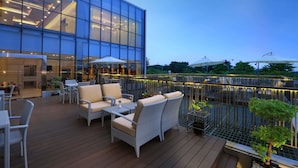 A modern outdoor seating area with wicker furniture and umbrellas, overlooking a building with large glass windows. - Harper Cikarang by ASTON (Cikarang)