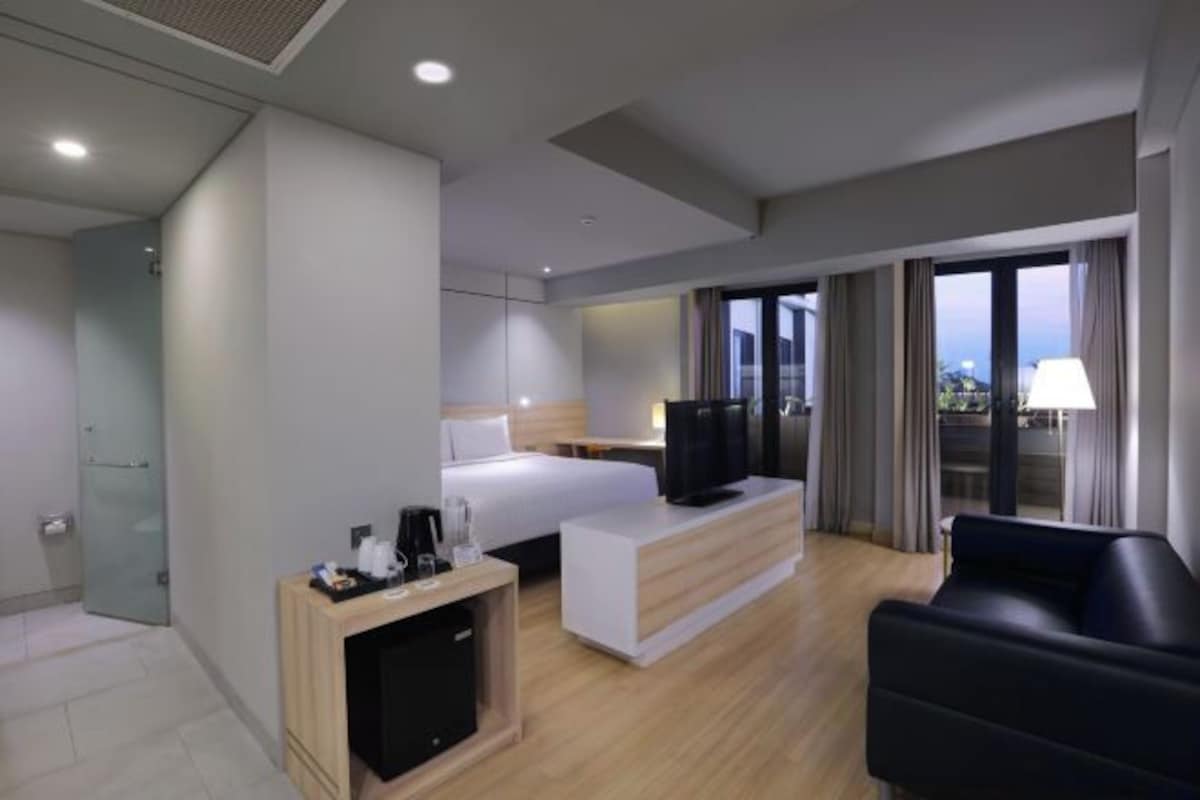A modern hotel room with a bed, a desk with a TV, a sofa, and a balcony with curtains.
