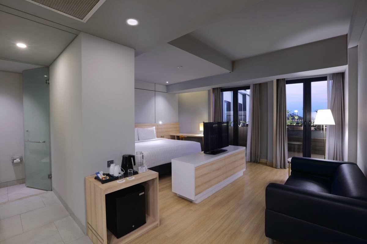 Una habitación de hotel moderna con cama, escritorio, televisor, sofá y balcón con cortinas.