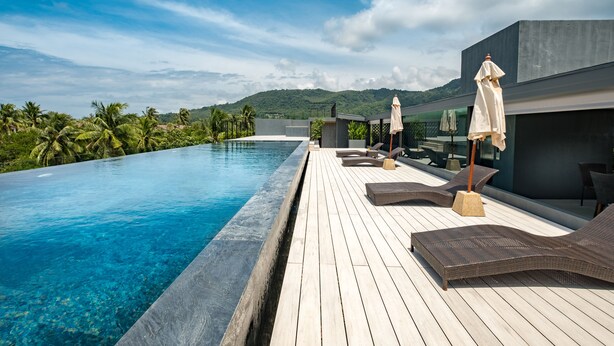 A rooftop infinity pool with wooden deck and lounge chairs, surrounded by lush greenery and mountains. - Kamala Resotel (Kamala)
