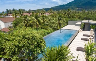 A rooftop infinity pool with a wooden deck and lounge chairs, surrounded by lush greenery and palm trees.