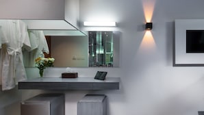 A modern bathroom with a large mirror, a vanity with a light, a small stool, and a towel rack. - Kamala Resotel (Kamala)
