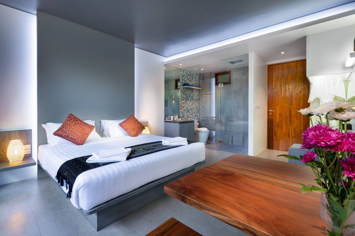 A modern hotel room with a large bed, a wooden coffee table, and a bathroom visible through a glass door.