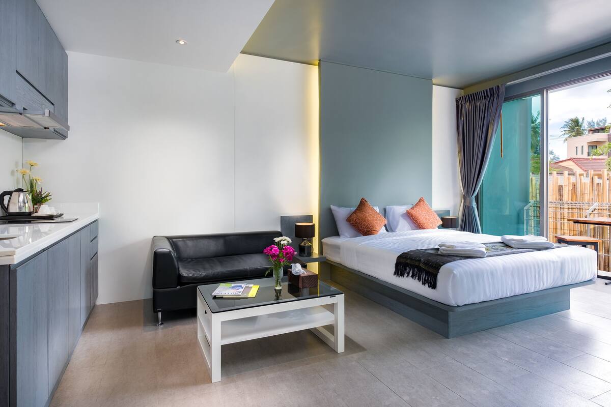 A modern hotel room with a bed, a sofa, a coffee table, and a kitchen area.