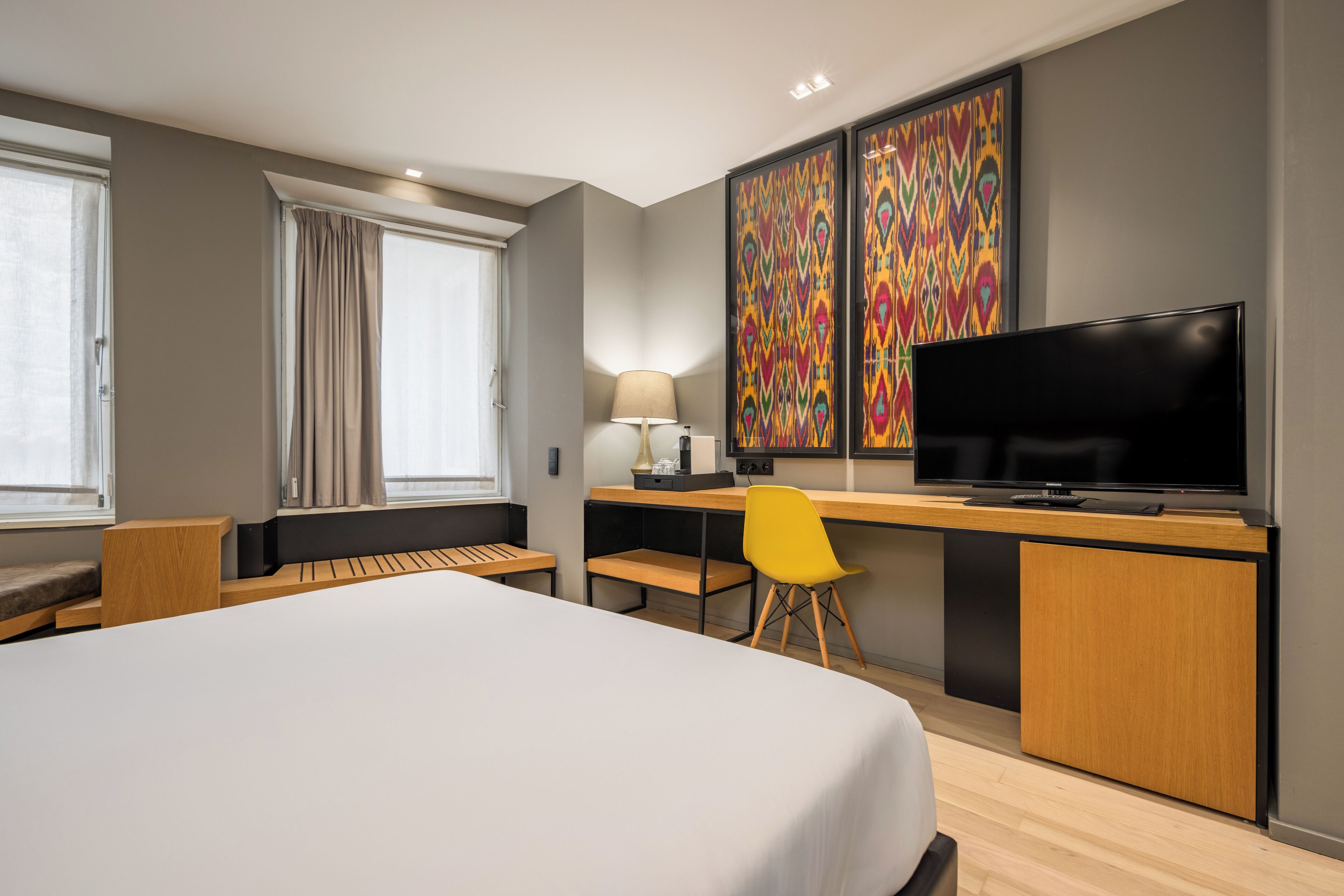 A modern hotel room with a large bed, a desk with a television, a chair, and a desk lamp.