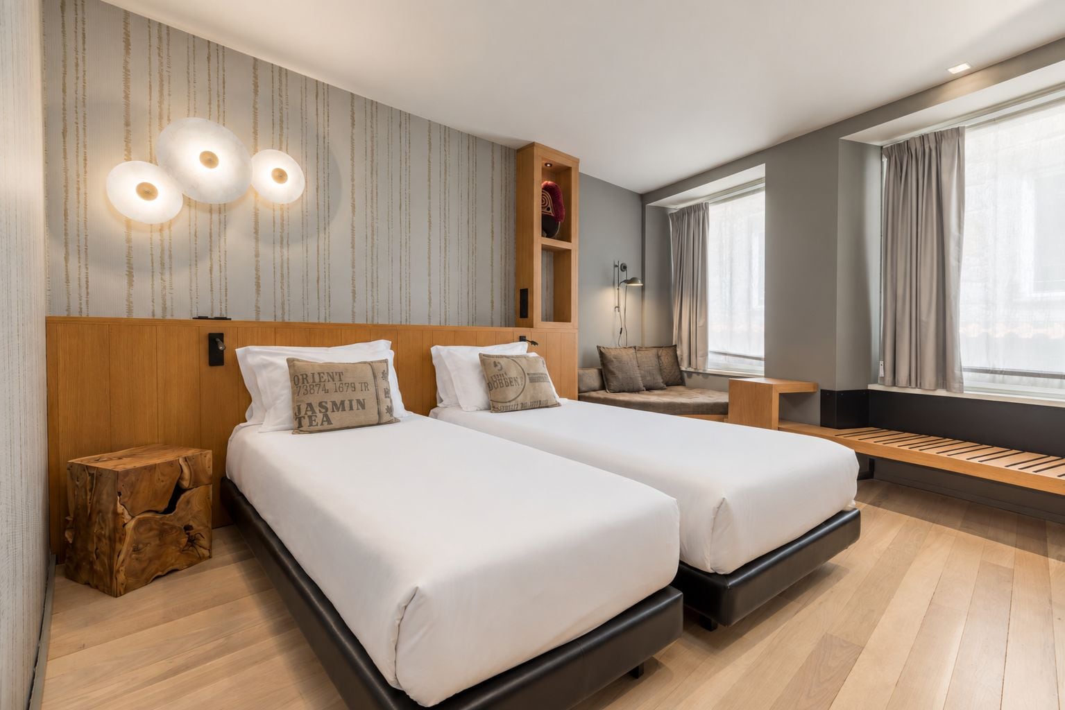 Habitación de hotel con dos camas, cabecera de madera, un taburete pequeño de madera, un banco y una ventana con cortinas.
