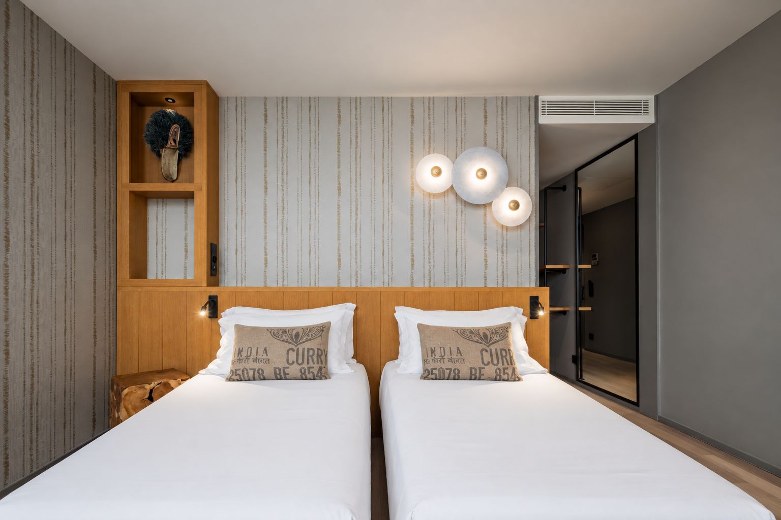 A hotel room with two beds, a wooden headboard, a wall-mounted shelf with a decorative item, and a wall-mounted light fixture.