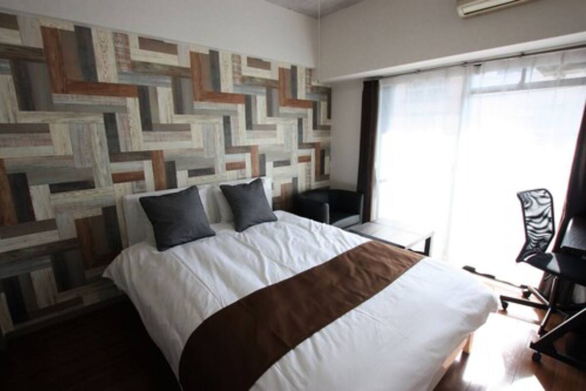 A hotel room with a large bed, a desk, and a chair. There is a window with curtains, and a wall with a geometric pattern.