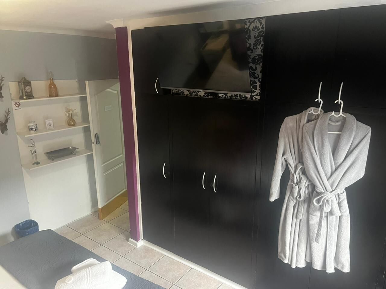 Une salle de bain avec un grand miroir, une armoire noire, une étagère avec des articles de toilette et deux peignoirs suspendus à des crochets.