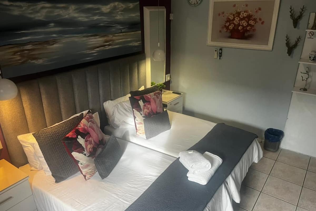A hotel room with a bed, a bedside table, a nightstand, a mirror, a clock, and a wall-mounted shelf with decorative items.