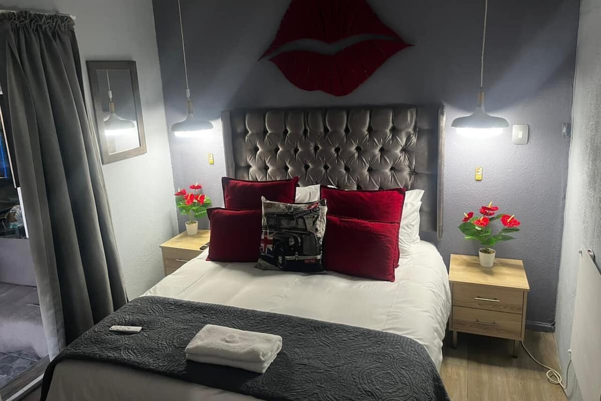 A bedroom with a tufted headboard, a large red lips artwork, two bedside tables with red and white pillows, and a bedside plant.