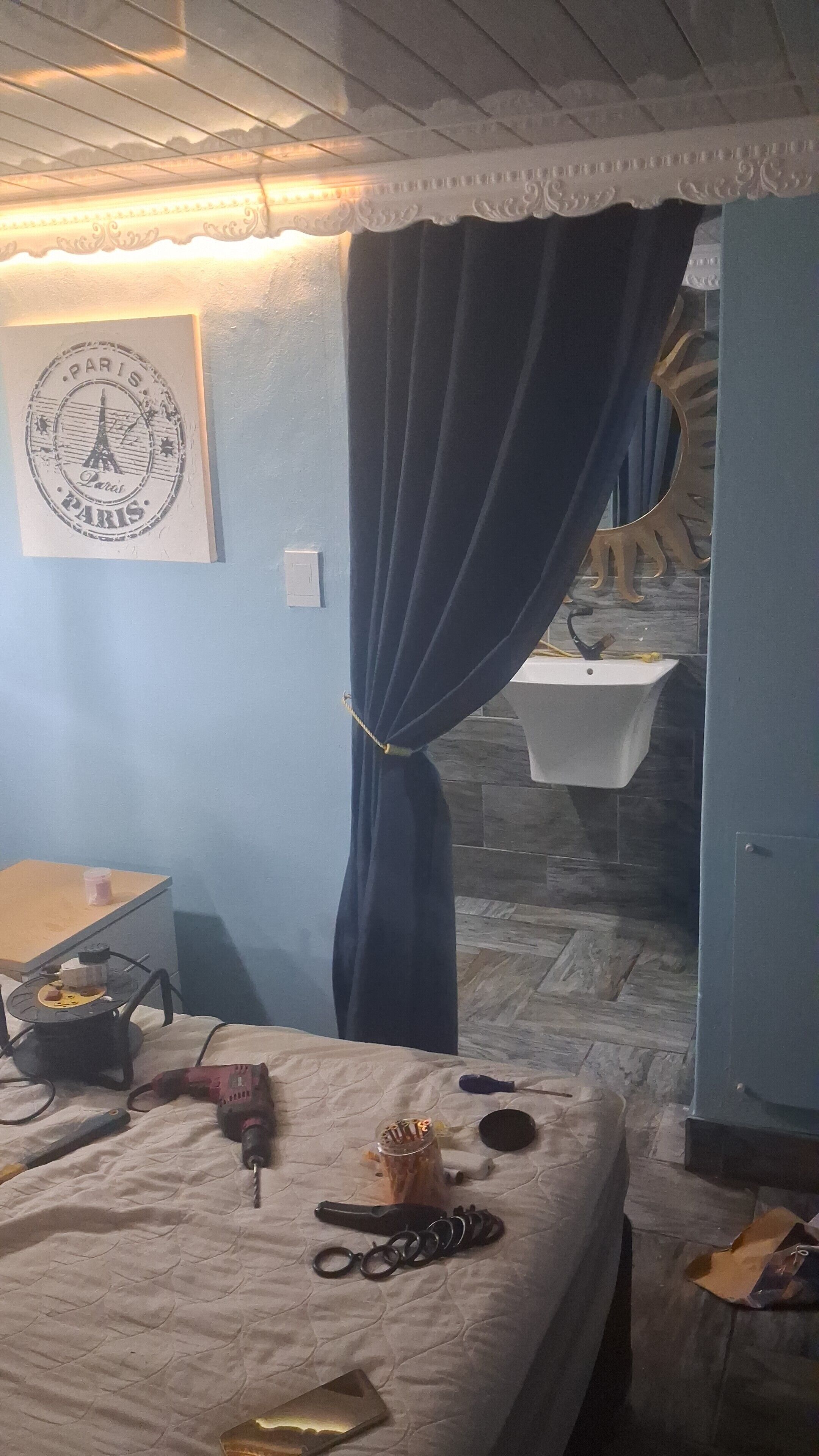 Een slaapkamer met een bed, een bureau, een aan de muur bevestigd schilderij, een badkamer met een wastafel en spiegel, en een blauw gordijn.