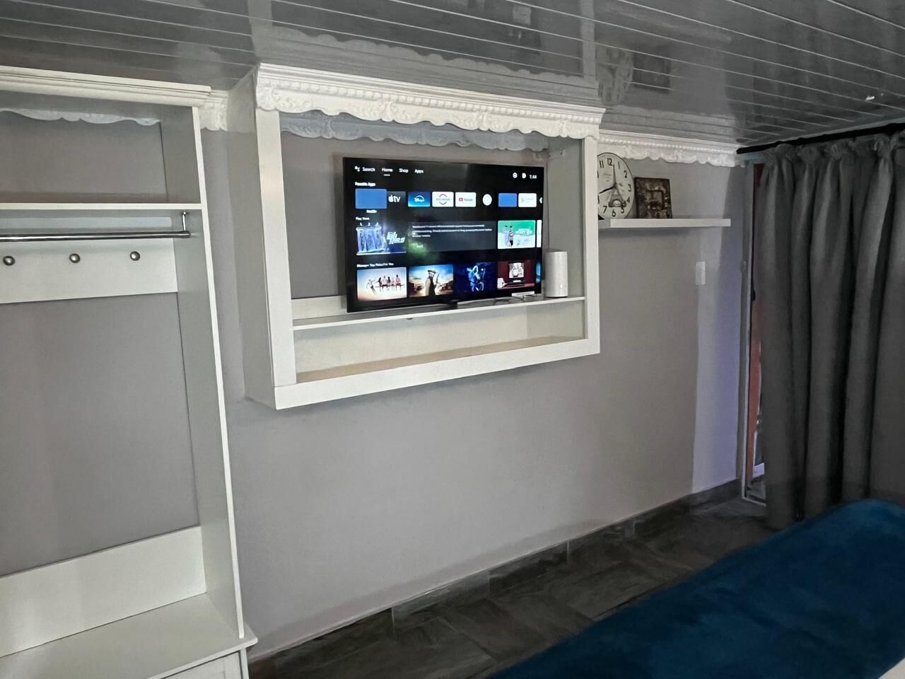 Ein Zimmer mit Fernseher an der Wand, einem Bett, einem Kleiderschrank und einem Fenster mit Vorhängen.
