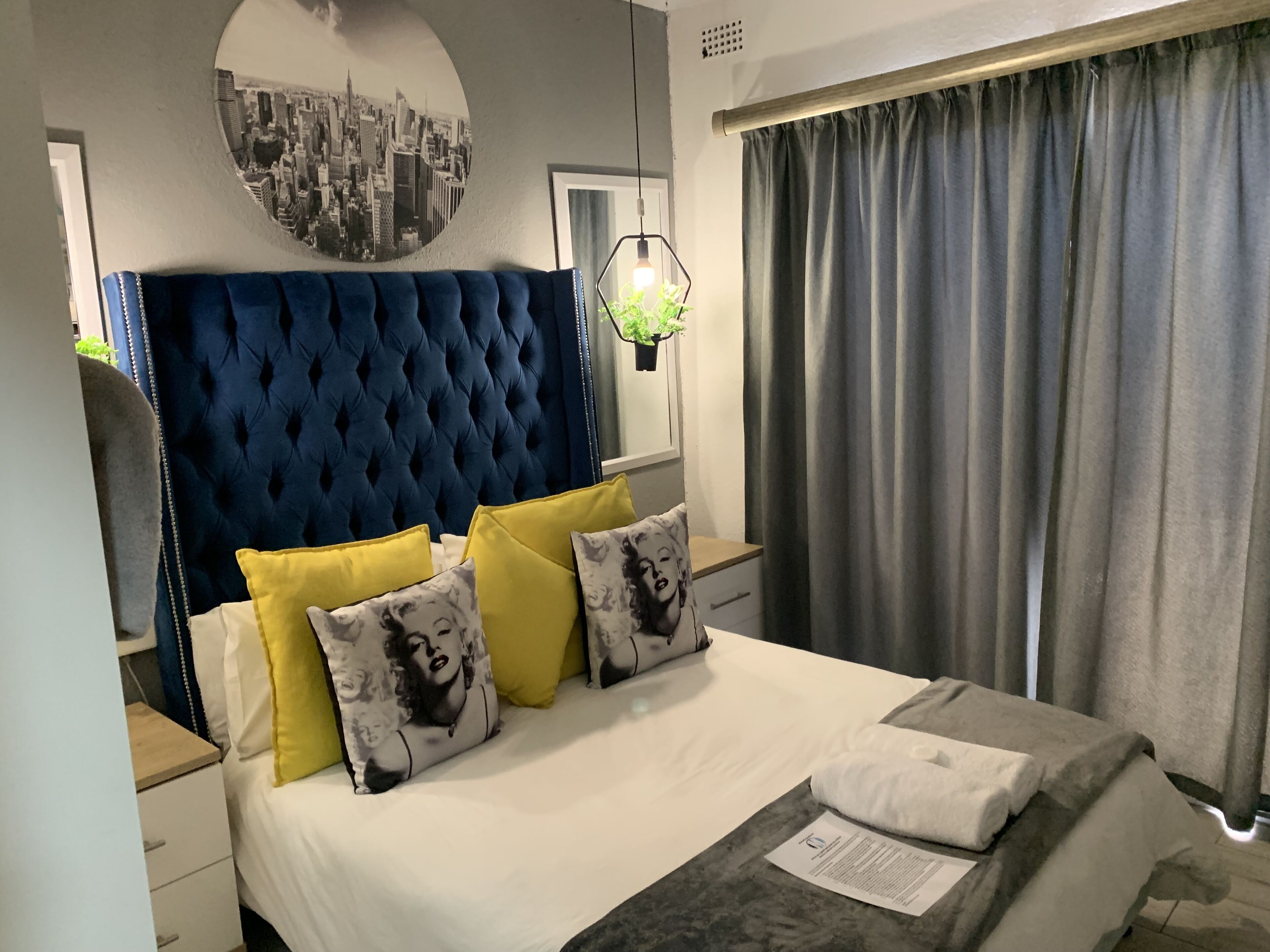 A neatly made bed with a blue headboard, yellow and patterned pillows, a white comforter, and a grey throw.