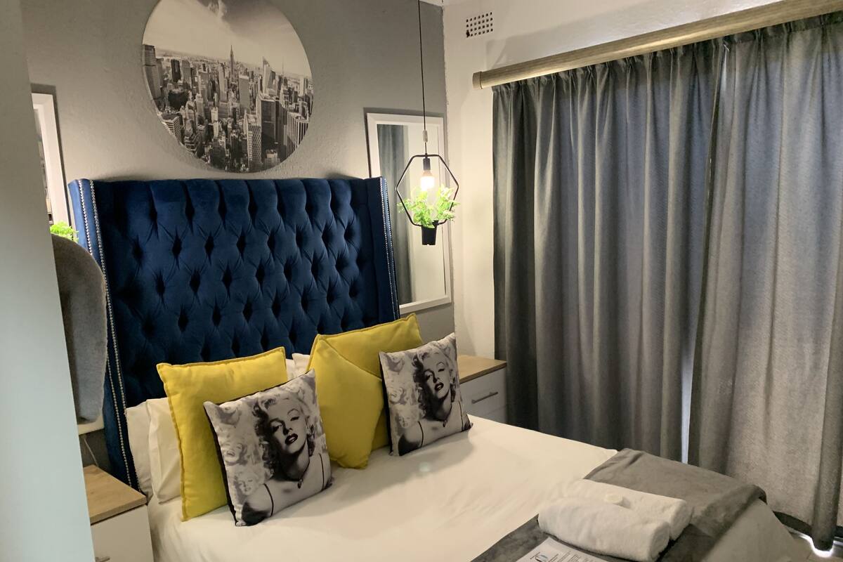 A neatly made bed with a blue headboard, yellow and patterned pillows, a white comforter, and a grey throw.