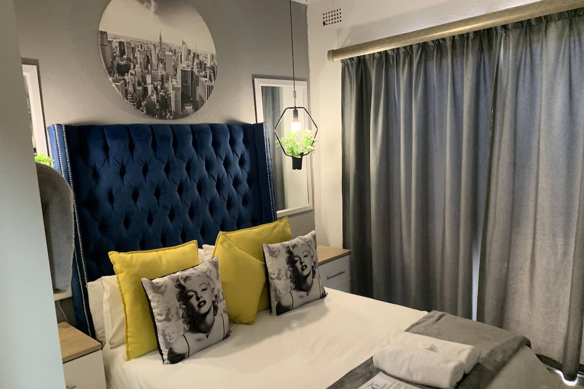 A neatly made bed with a blue headboard, yellow and patterned pillows, a white comforter, and a grey throw.