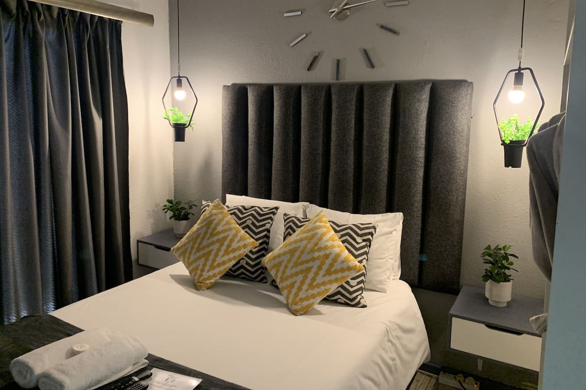 A modern hotel room with a large bed, two bedside tables, a wall clock, and hanging plants.