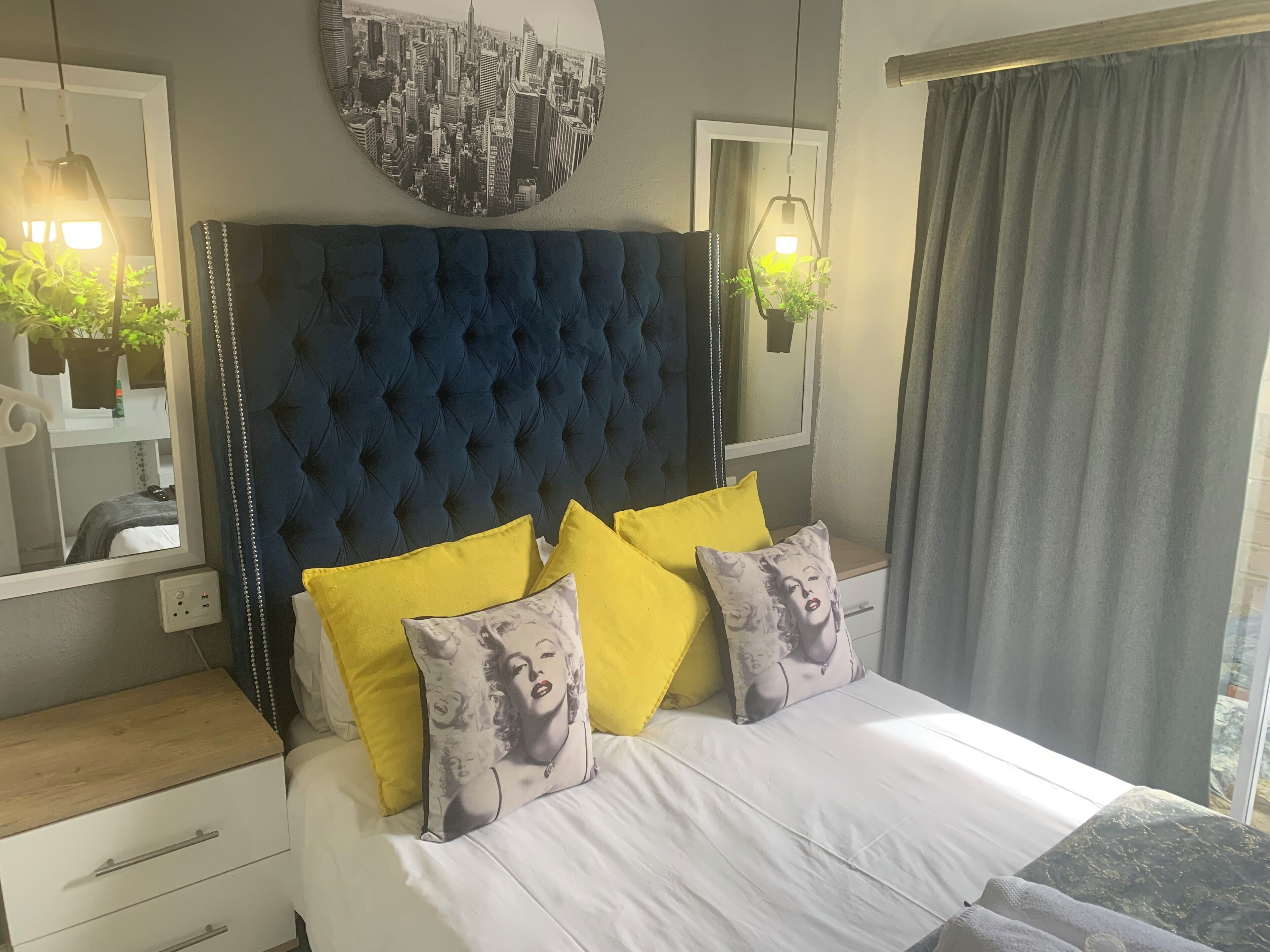 A bedroom with a tufted headboard, a bed with white bedding and multiple pillows, a bedside table, a mirror, and a cityscape artwork on the wall.