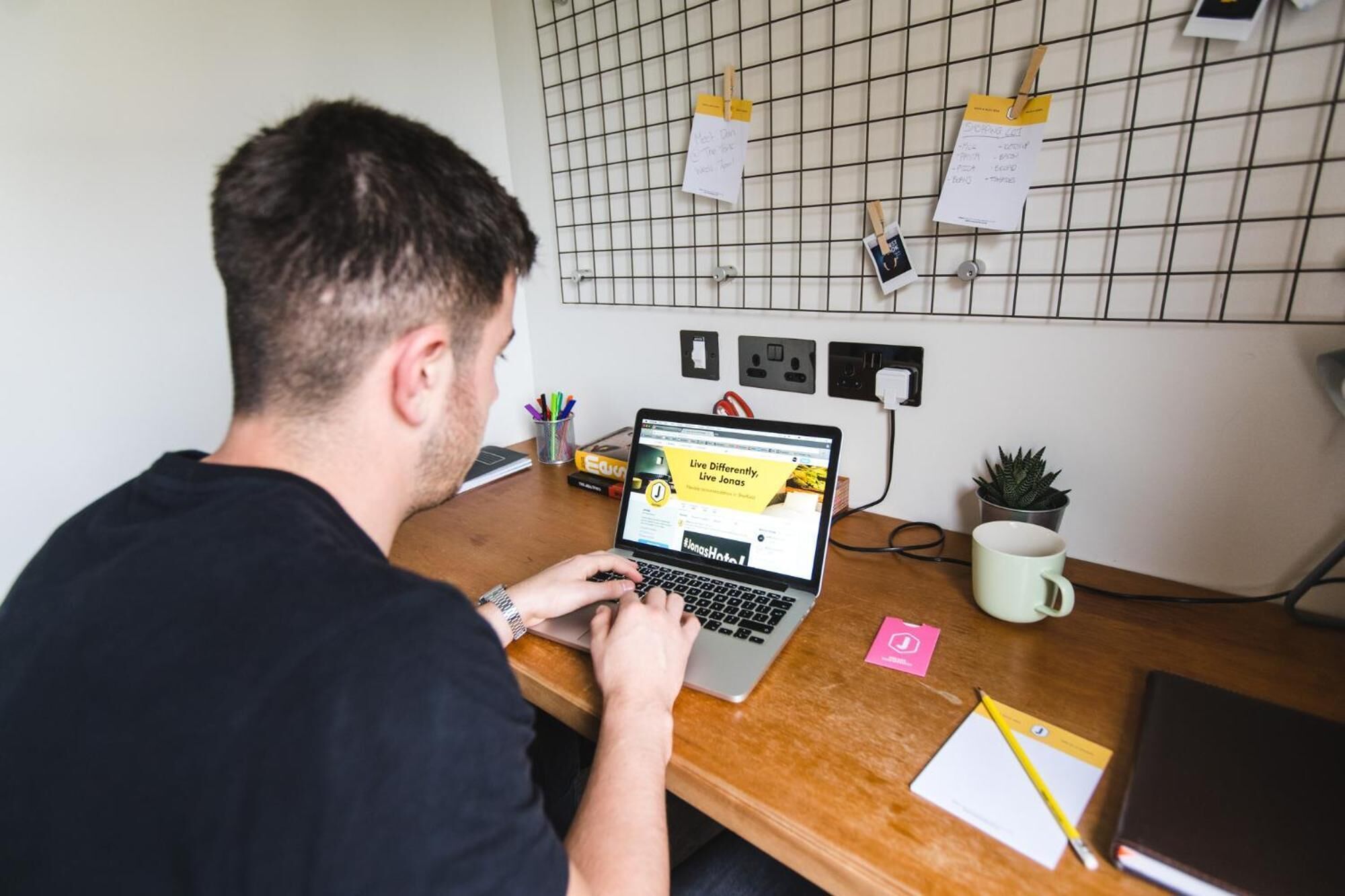 Une personne travaille à un bureau en bois, devant un ordinateur portable, avec une tasse et une petite plante.