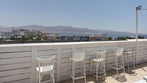 Una terraza con sillas y mesa de metal blanco, con vistas a un paisaje costero y montañas al fondo.