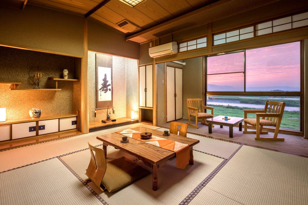 A traditional Japanese room with tatami flooring, a low wooden table, and chairs. There is a large window with a view of the sea.