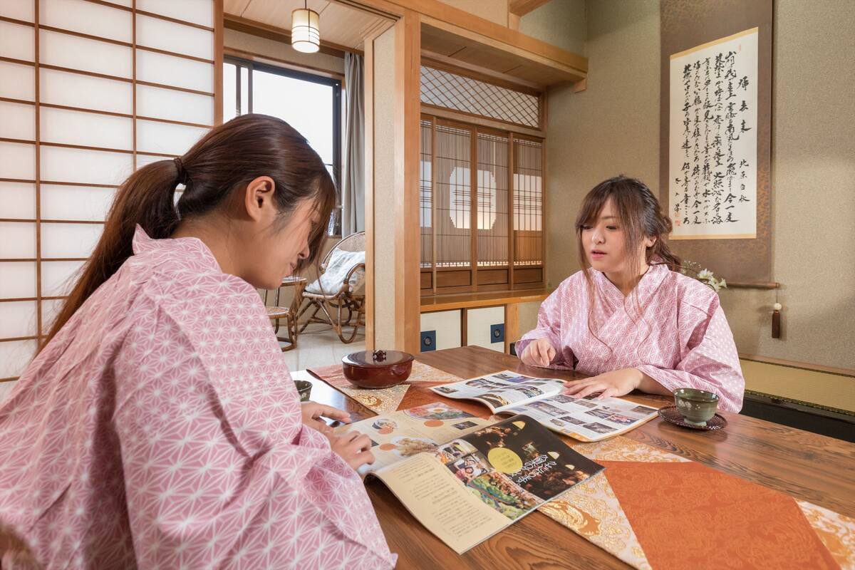 Two people in traditional attire sitting at a wooden table with magazines and a teapot.