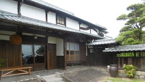 A traditional Japanese building with a tiled roof, wooden doors, and a stone pathway leading to a wooden bench. - Yumoto Kobayashi (Kurume)