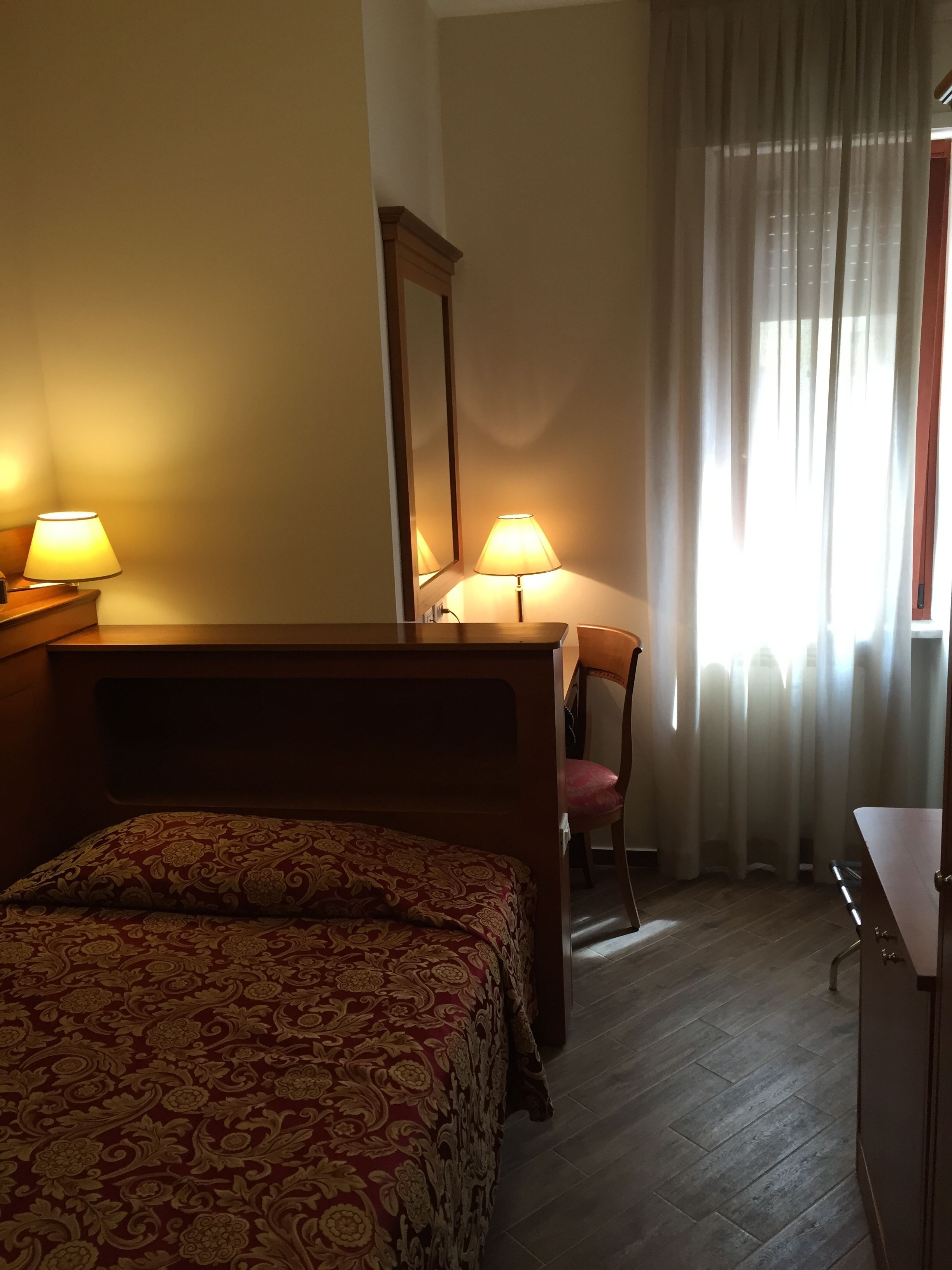 A hotel room with a bed, a desk with a chair, a mirror, and a window with curtains.
