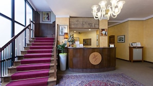 A hotel reception area with a wooden reception desk, a large potted plant, a red carpeted staircase, and a patterned rug. - Hotel Excelsior (Pavia)