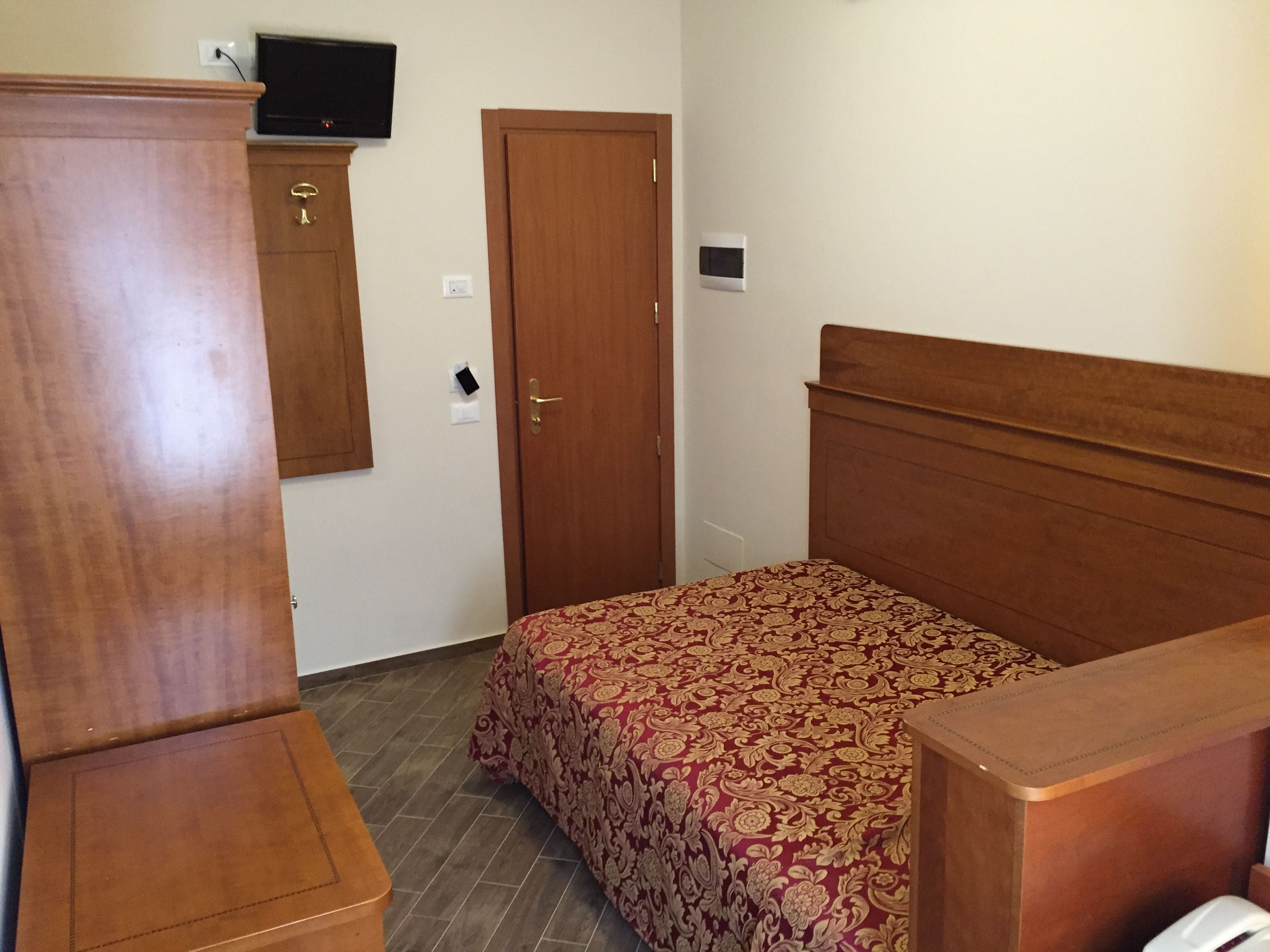 A bedroom with a wooden bed, a wardrobe, a television on the wall, and a thermostat.