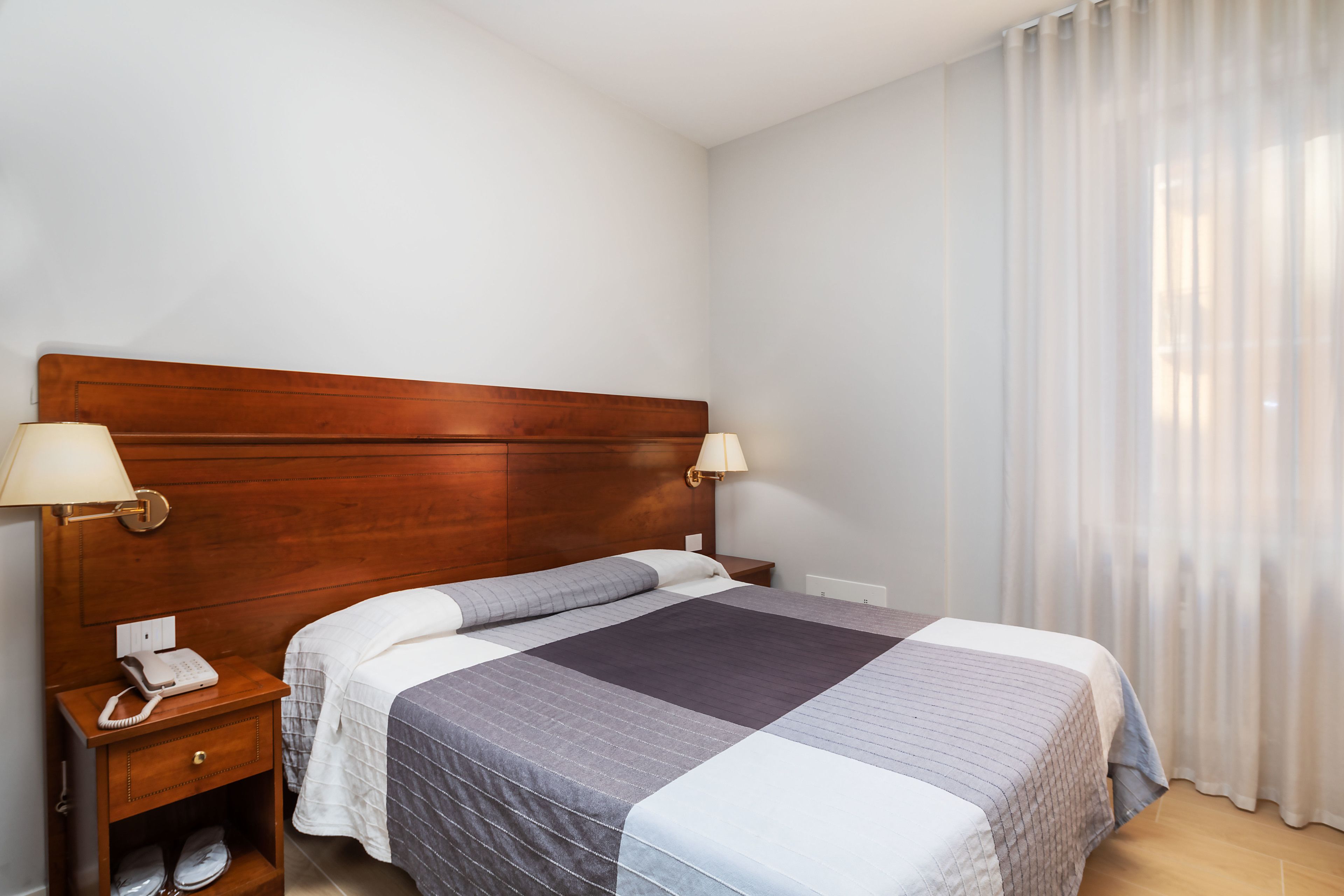 Habitación de hotel con cabecera de madera, dos lámparas de noche, un teléfono, un cajón pequeño y una cama con colcha a rayas.