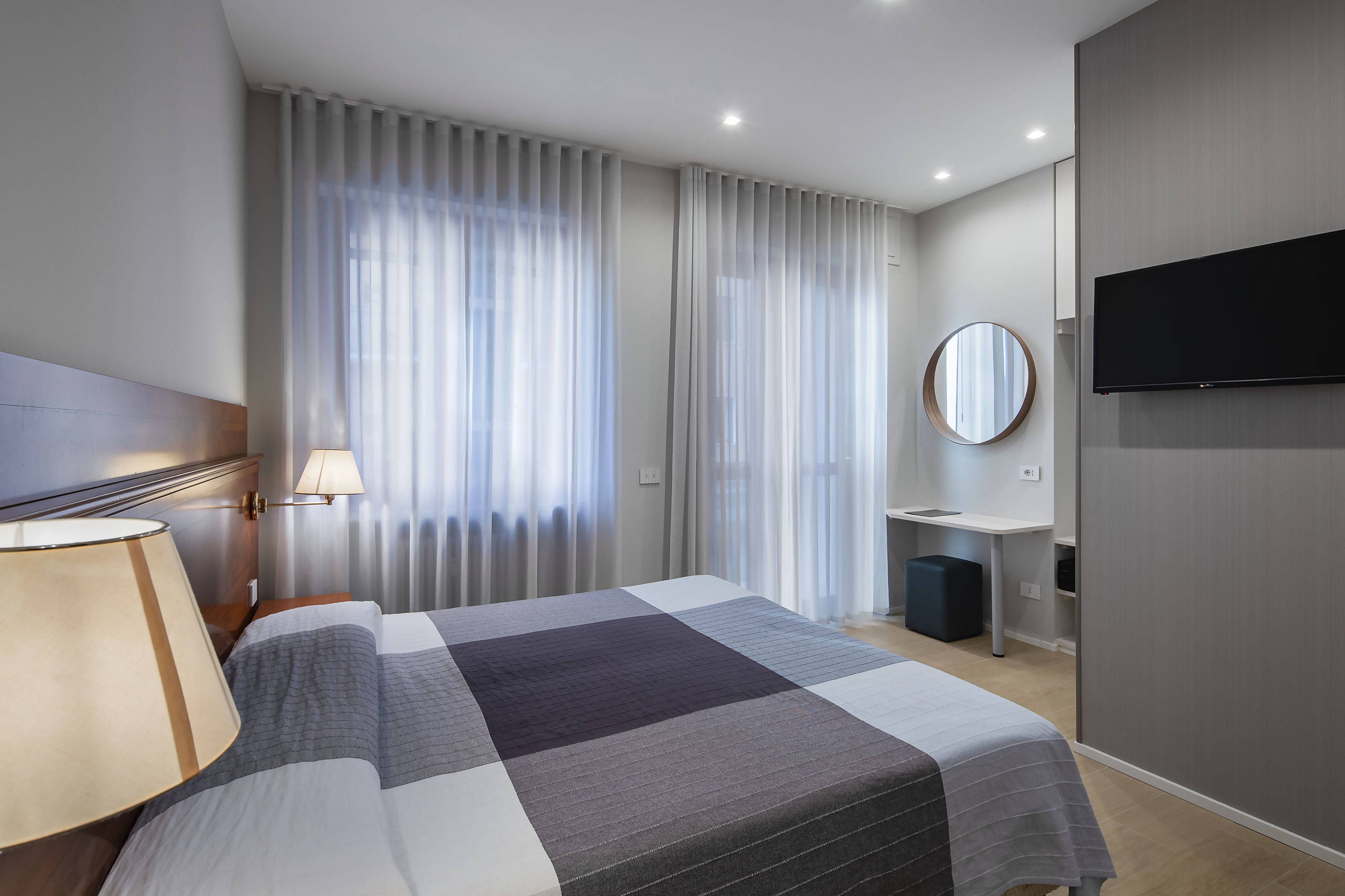 A modern hotel room with a large bed, a bedside lamp, a flat-screen TV, a round mirror, and a small desk with a chair.
