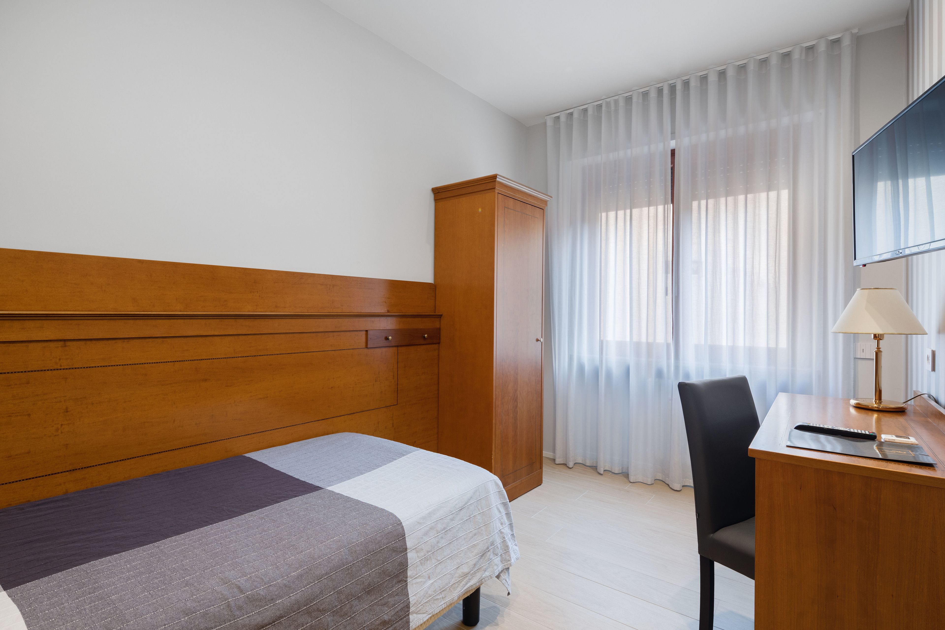 Ett hotellrum med en säng, ett skrivbord, en stol, en garderob och ett fönster med gardiner.