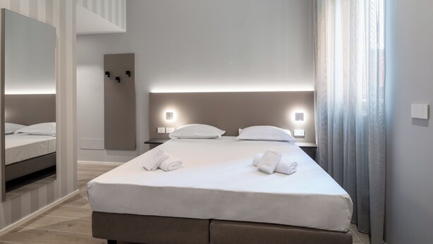 A modern hotel room with a large bed, bedside tables, a mirror, and a view of the city through the window. - Hotel Excelsior (Pavia)