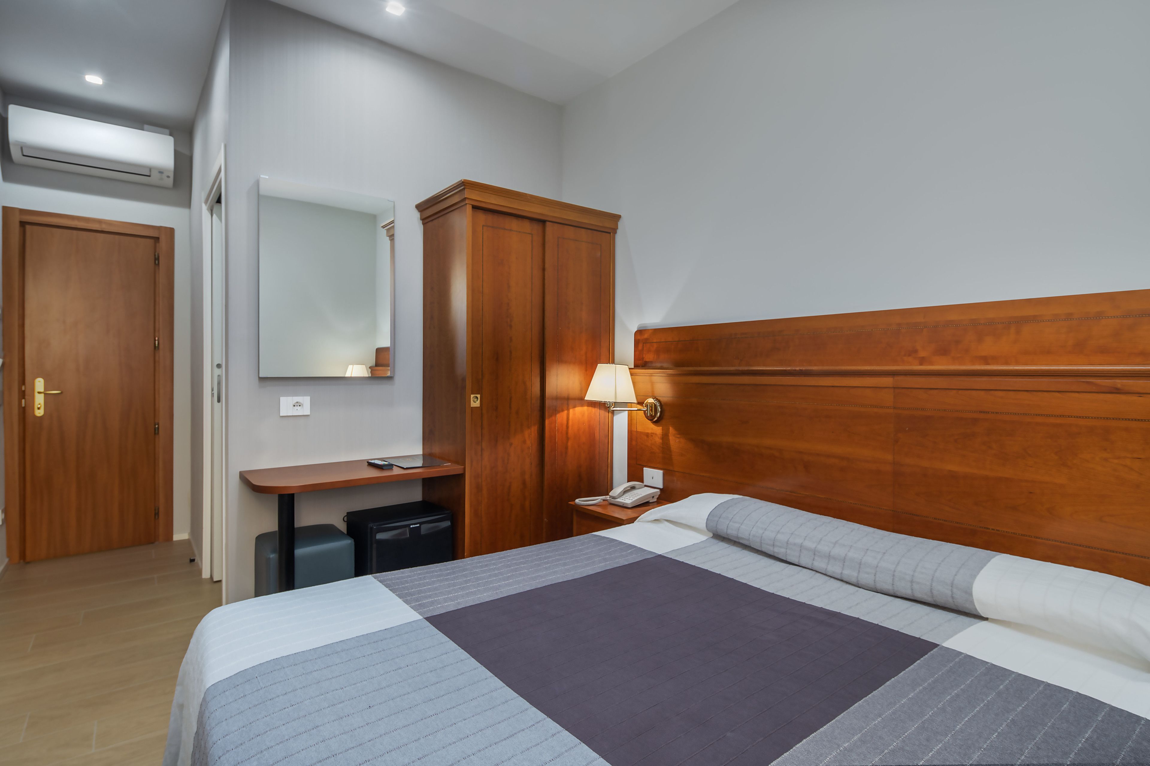 A hotel room with a wooden headboard, a bed with a striped bedspread, a desk with a lamp, a telephone, and a wardrobe.