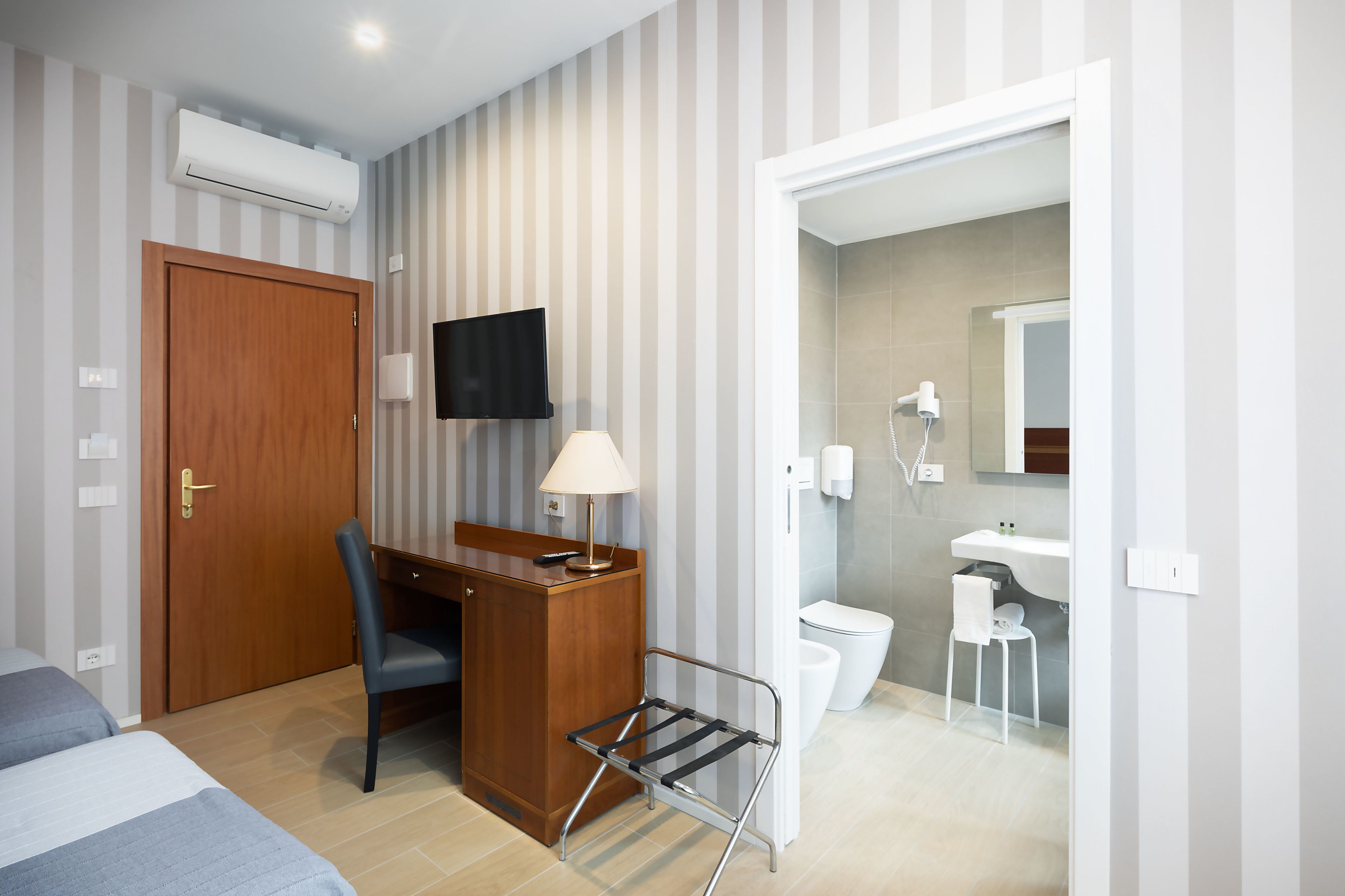 Et hotelværelse med seng, skrivebord, stol og et lille bord med lampe. Der er et TV ophængt på væggen, et badeværelse med toilet og håndvask, samt en bruser.