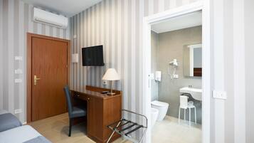 Et hotelværelse med seng, skrivebord, stol og et lille bord med lampe. Der er et TV ophængt på væggen, et badeværelse med toilet og håndvask, samt en bruser.