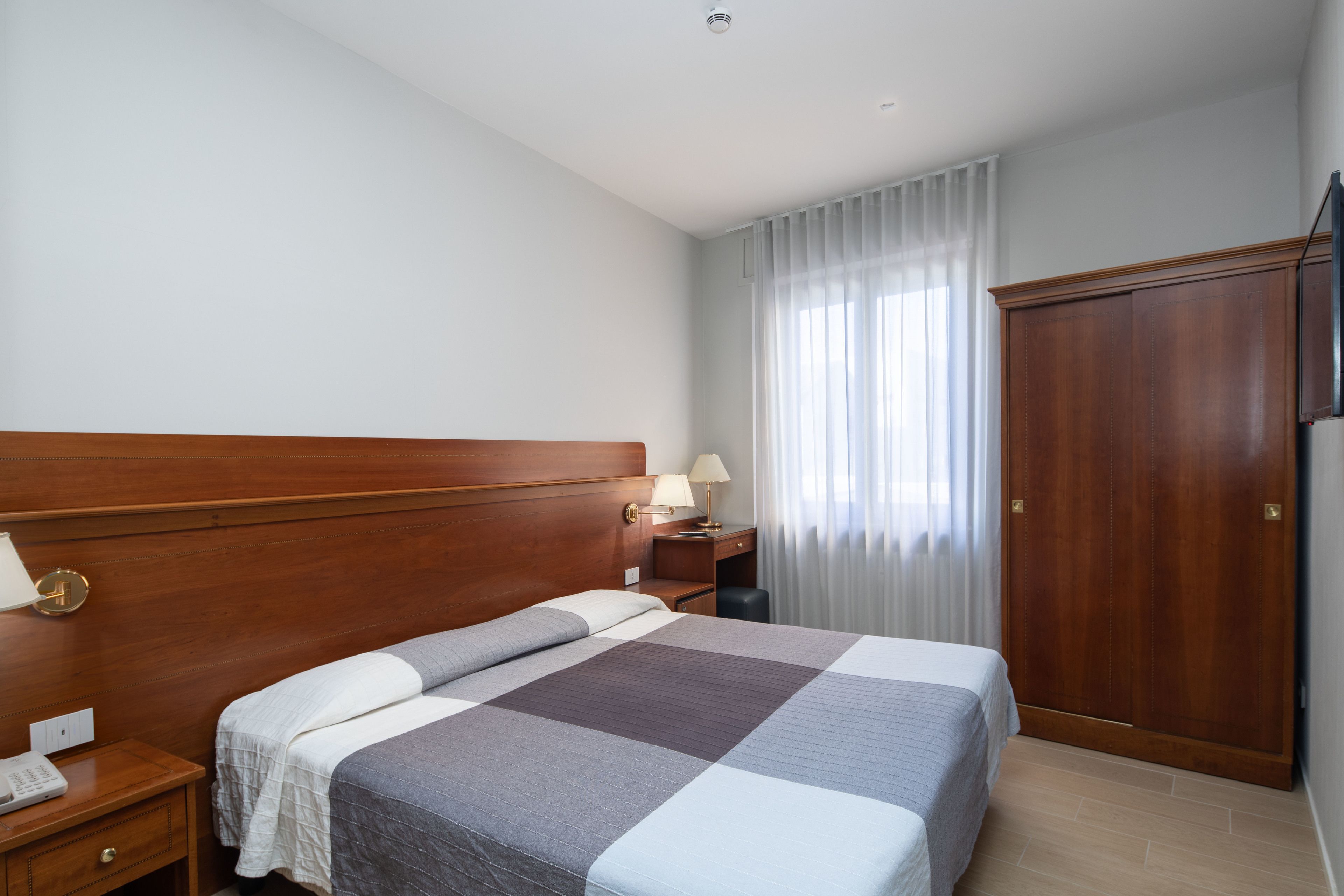Ett hotellrum med en sänggavel i trä, en säng med ett rutigt överkast, ett sängbord med en lampa, ett litet bord, en garderob i trä och ett fönster med tunna, genomskinliga gardiner.