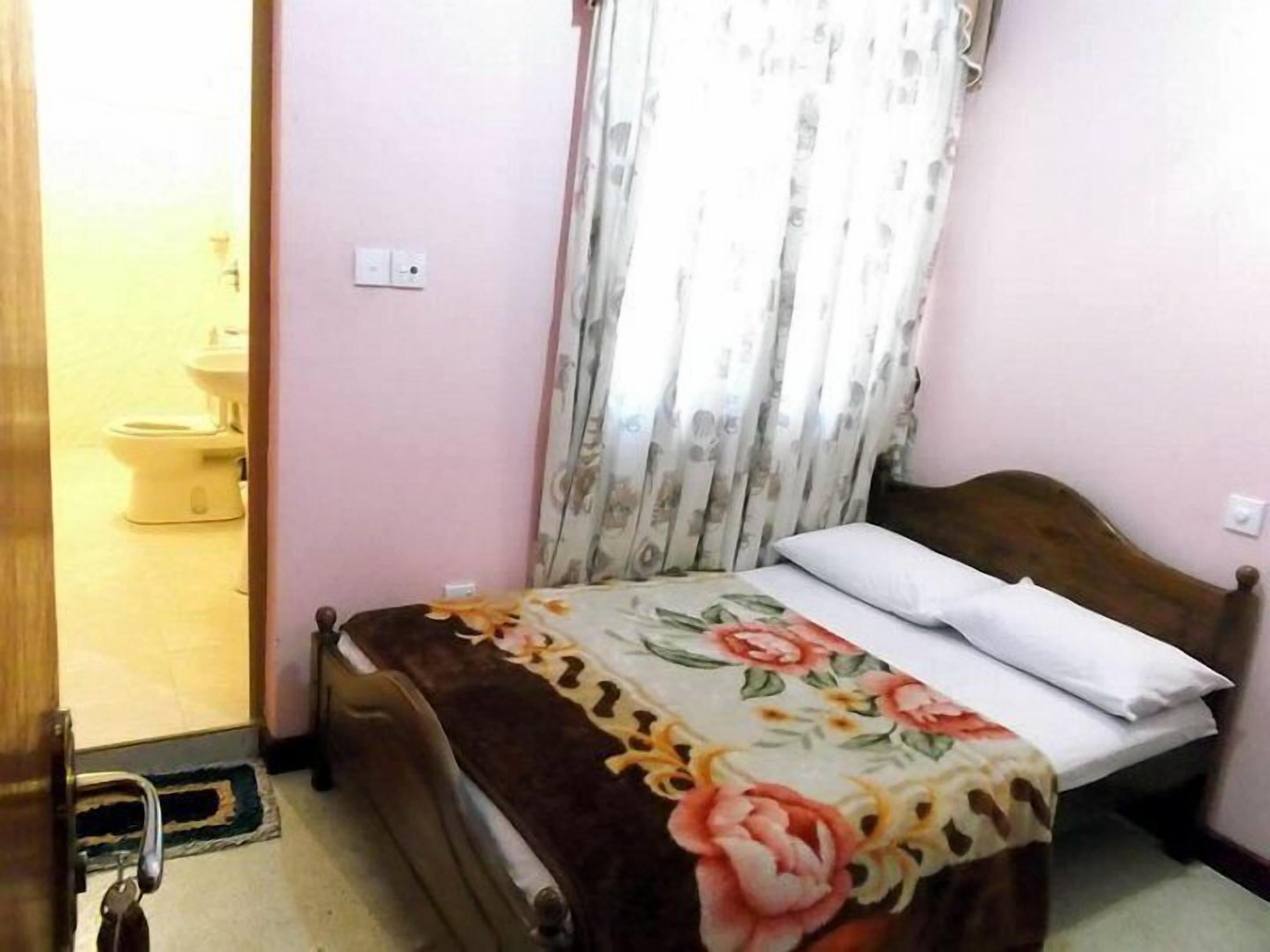 A bedroom with a bed, a wooden headboard, a floral bedspread, a white pillow, and a door leading to a bathroom with a toilet.
