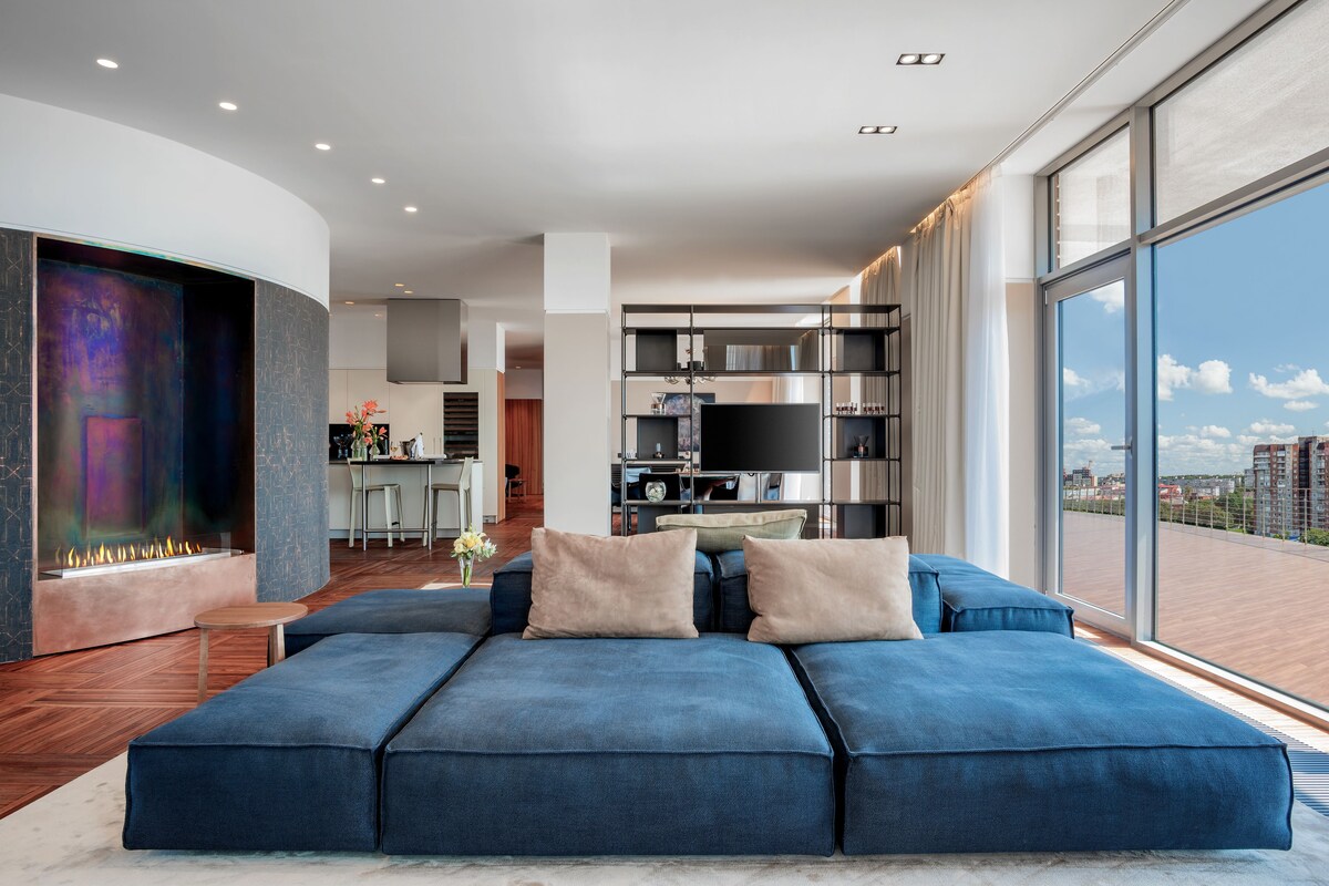 Un salon moderne avec un canapé d’angle bleu, une cheminée et une grande fenêtre donnant sur la ville.