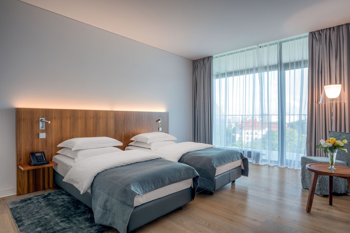 Une chambre d’hôtel moderne avec deux lits, une tête de lit en bois, un téléphone sur la table de chevet, une petite table avec un vase de fleurs et une grande fenêtre avec des rideaux.