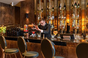 Un espace bar avec un barman préparant une boisson, des tabourets de bar, et une variété de bouteilles et de verres disposés sur des étagères.