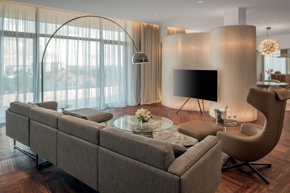Un salon moderne avec un canapé d’angle, une table basse en verre, un fauteuil aux lignes courbes et un téléviseur à écran plat.