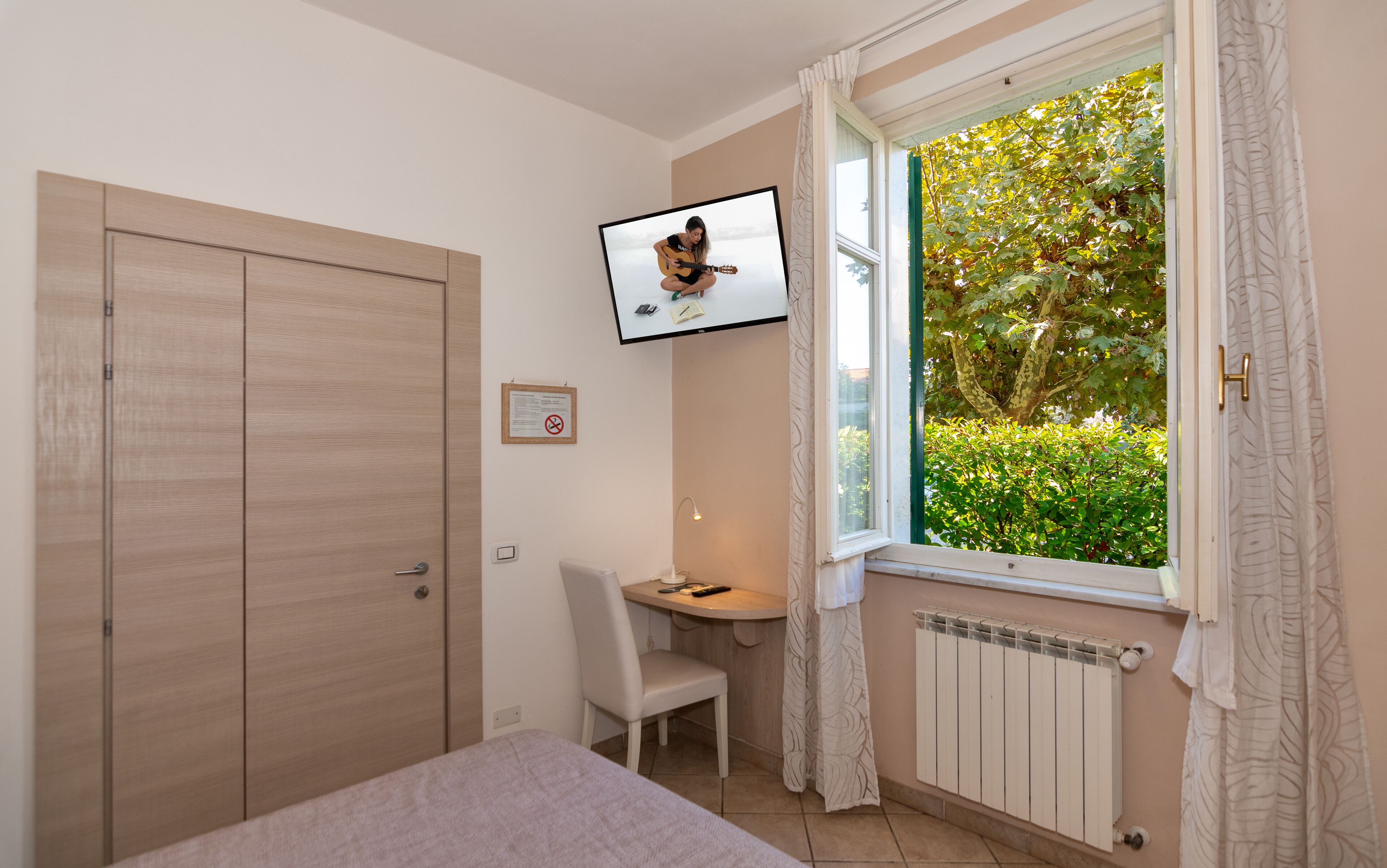 A bedroom with a bed, a desk, a chair, a TV mounted on the wall, and a window with curtains.