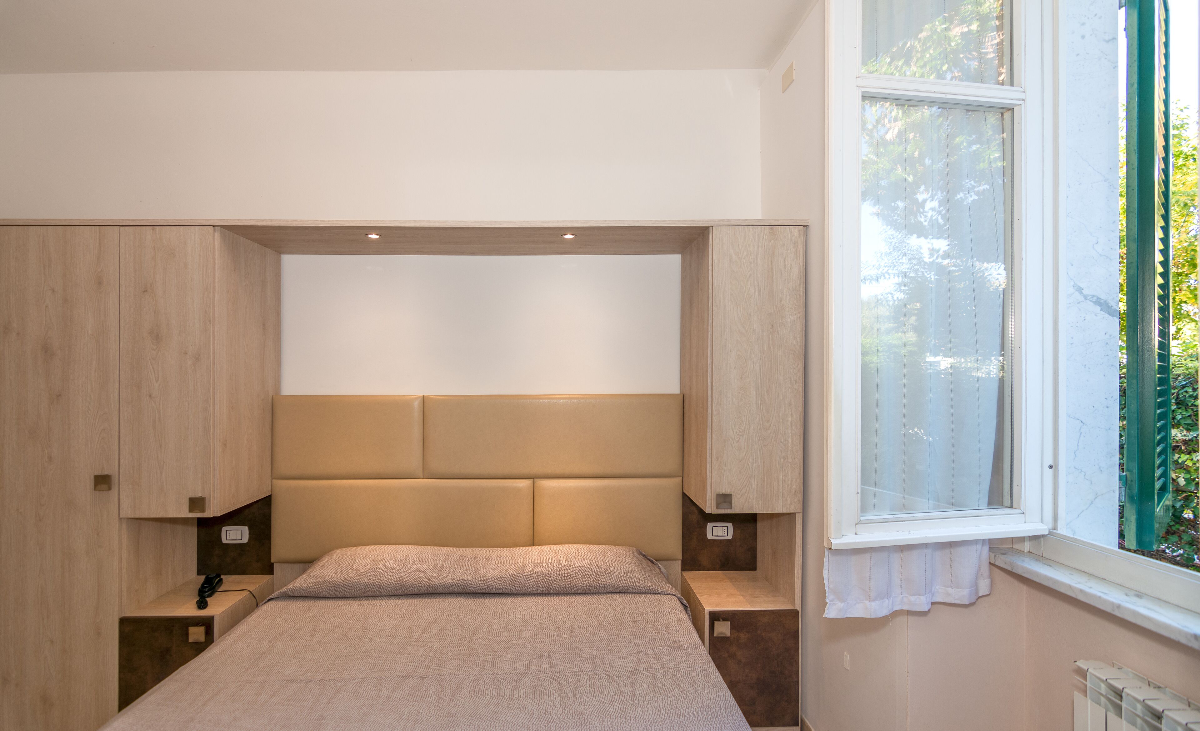 A bedroom with a large bed, wooden headboard, bedside tables, and a window with a view of greenery.