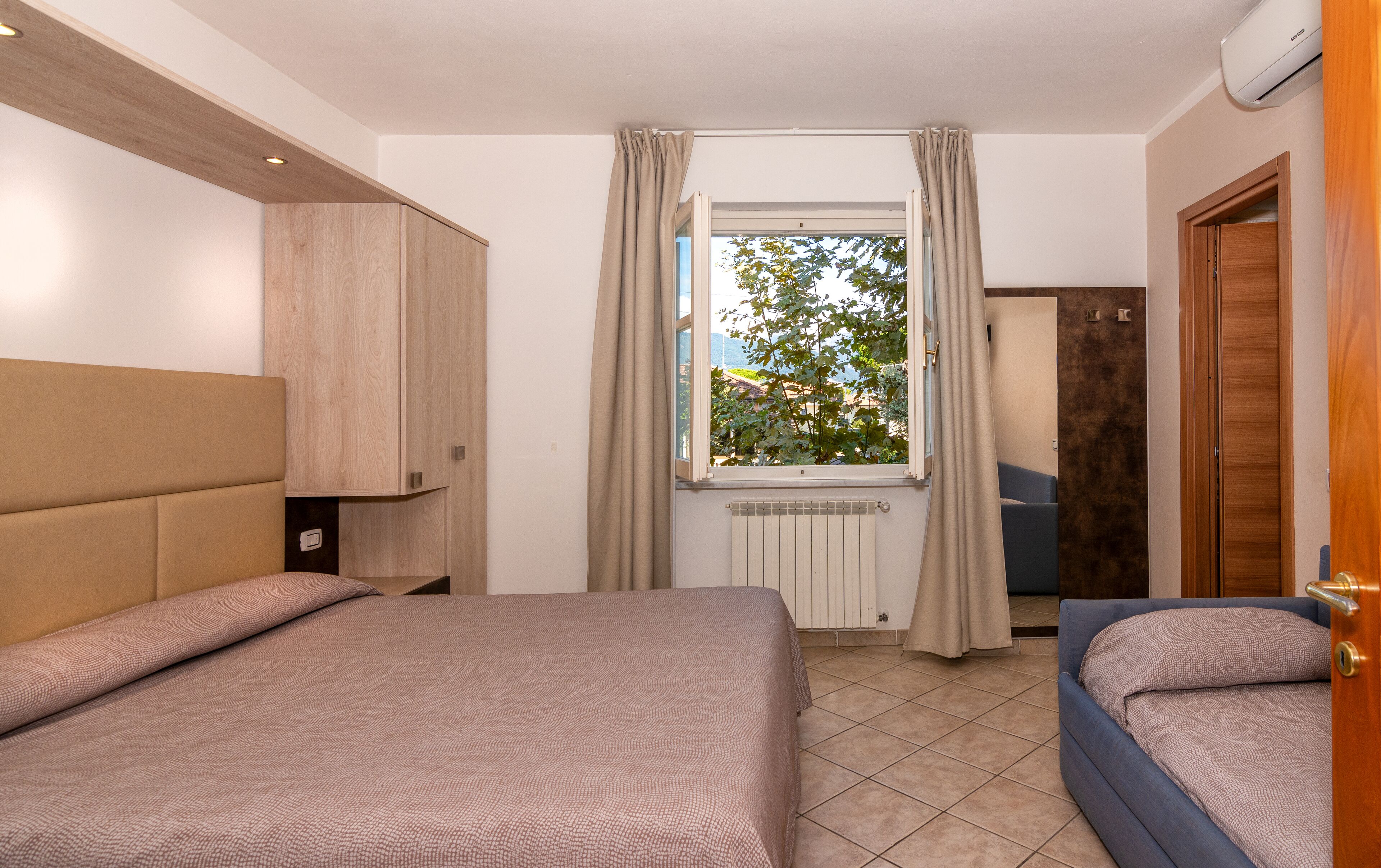 A hotel room with two beds, a window with curtains, a radiator, and a view of trees and buildings outside.