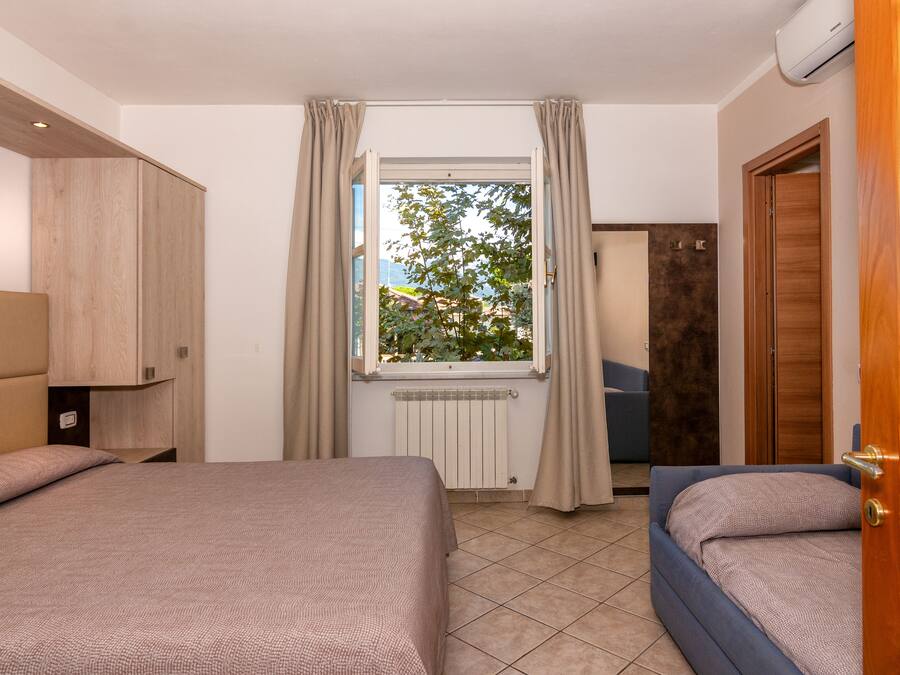 A hotel room with two beds, a window with curtains, a radiator, and a view of trees and buildings outside.